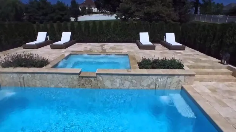 A luxurious outdoor pool area with clear blue water features a raised hot tub, surrounded by four white lounge chairs on a stone patio—perfect for relaxing or filming stunning pool videos, all bordered by tall green hedges. Luxury Pool