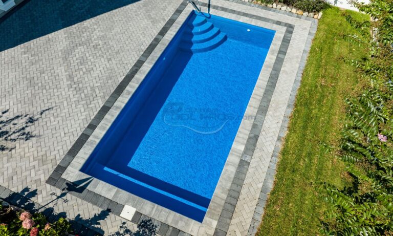Aerial view of a modern rectangular swimming pool with clear blue water, surrounded by gray stone paving and a well-maintained green lawn. Ideal for outdoor relaxation and recreation, luxury pool builder.