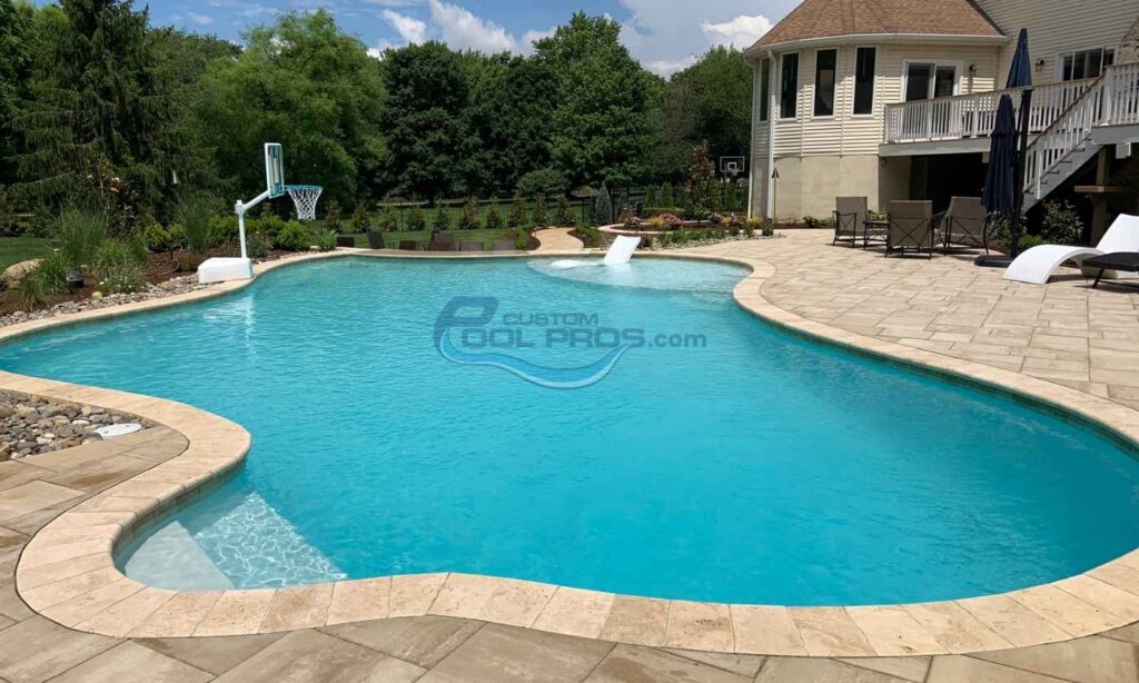 Backyard swimming pool with clear blue water, surrounded by a stone patio, a basketball hoop, and lush greenery in the background, perfect for outdoor relaxation and recreation, custom inground pools.