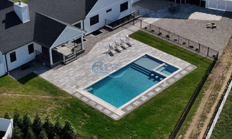 Aerial view of a modern backyard featuring a rectangular swimming pool surrounded by a stone patio. Lounge chairs are positioned nearby, and a covered porch is attached to the house. The area is enclosed with a fence, showcasing a well-manicured lawn and gravel landscaping, inground pools
