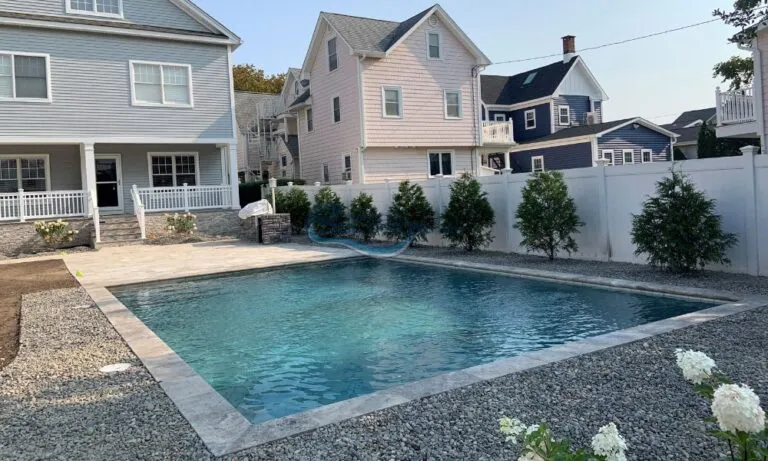 Luxury pools, modern backyard featuring a rectangular swimming pool surrounded by gravel and landscaped greenery, with a two-story house in the background. The scene showcases a relaxing outdoor space ideal for leisure and entertainment, pool contractors